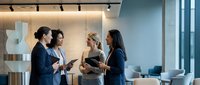 Young diverse female professionals in business casual attire having collaborative discussion in modern office environment with natural lighting and contemporary workspace design.