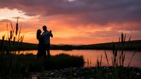 Romantic couple silhouette embracing by lake at golden sunset with cattails and reeds, peaceful evening landscape for love and relationship concepts.