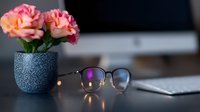 Pink roses in speckled blue pot with eyeglasses on desk surface, soft bokeh lights create cozy home office atmosphere. Modern workspace styling.