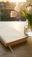 White tulips in glass vase beside closed book on wooden table with warm golden sunlight streaming through window creating peaceful morning atmosphere.