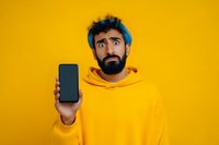 Surprised bearded man in yellow hoodie holding smartphone with blank screen on bright yellow background for mobile app presentation and technology advertising.