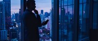Silhouette of businessman using smartphone by office window overlooking illuminated city skyline at dusk for corporate communication and urban lifestyle concepts.