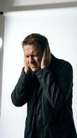 Stressed middle-aged Caucasian man covering ears with hands, eyes closed, expressing frustration and anxiety, wearing black jacket on light background.