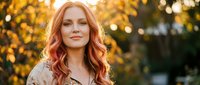 Beautiful redhead woman with wavy hair smiling outdoors during golden hour sunset with warm bokeh lights and autumn foliage background for lifestyle photography.