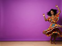 Young woman with dark skin tone and voluminous curly hair wearing vibrant multicolored patterned dress, dancing joyfully with arms raised against solid purple background with wooden floor.