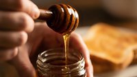 Unrecognizable person holding wooden honey dipper dripping golden honey into glass jar with bread in background for food and cooking content.
