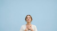 Young woman with brown hair and fair complexion, wearing a light beige cardigan over a white top, hand placed on chest, smiling warmly against a soft blue background for portrait and wellness use.