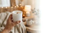 Woman holding lit scented candle in cozy bedroom with warm string lights and soft furnishings, creating relaxing ambiance for wellness and home decor.