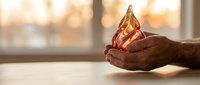 Young adult male with dark skin tone holding a glowing golden teardrop or flame-shaped object in cupped hands, symbolizing hope, spirituality, or precious value in warm indoor lighting.