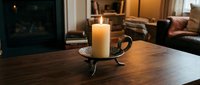 Lit pillar candle in ornate metal holder on wooden table, cozy living room with fireplace in background, warm ambient light.