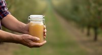 Male farmer holding glass jar of fresh apple cider or juice outdoors in orchard. Harvest season beverage production and organic farming concept for agricultural business.