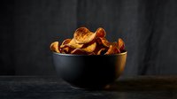 Crispy golden potato chips in black ceramic bowl on dark wooden table against moody background for snack food advertising and culinary photography.