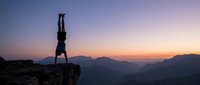 Athletic man performing handstand on mountain cliff edge during golden hour sunset with dramatic sky and valley landscape below for adventure sports content.