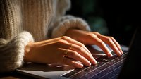 Female hands typing on laptop keyboard wearing cozy beige sweater and gold ring, soft natural lighting creates warm atmosphere for remote work and productivity.