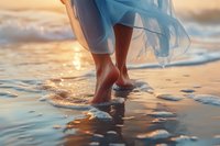 Barefoot woman in flowing blue dress walking on wet beach sand during golden sunset, waves gently washing over feet creating reflective water surface.