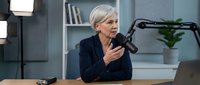 Professional mature Caucasian businesswoman recording podcast in modern office studio with microphone and laptop for content creation and broadcasting.