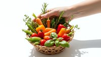 Hand reaching for fresh organic vegetables and herbs in wicker basket including tomatoes, peppers, rosemary and basil on white background for healthy cooking.