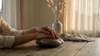Hands resting on smooth stone during meditation session with warm natural lighting and dried branches creating peaceful zen atmosphere for mindfulness practice.