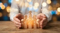 Hands protecting wooden family figures with glowing light, representing care, unity, insurance, support and community protection services.