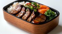 Grilled teriyaki glazed pork belly slices served in wooden bento box with steamed rice, fresh carrots and garnished with chopped green onions and parsley.