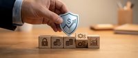 Business hand holding security shield over wooden blocks with financial icons representing data protection and risk management strategy.