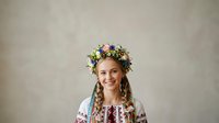 Young woman with long braided hair wearing traditional embroidered white blouse and colorful floral crown, smiling at camera against neutral background for cultural heritage and festive occasions.