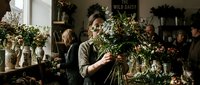 Female florist with dark hair arranging white and green flowers in studio workshop surrounded by botanical materials and floral design elements.