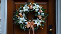Easter wreath with white lilies, pastel eggs, eucalyptus leaves and peach bow hanging on wooden door creates welcoming spring holiday decoration.