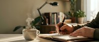 Person writing in notebook at cozy home office desk with coffee cup, lamp and books creating productive workspace atmosphere for journaling or work.