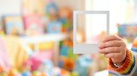 Child holding blank polaroid frame in playroom with colorful toys and decorations in soft focus background. Suitable for creative projects, memory concepts, and children's content.