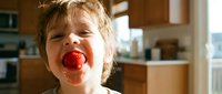 Young child with blonde curly hair eating red apple in bright kitchen, healthy snack and nutrition concept for families and children's wellness content.