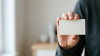 Man in dark sweater holding blank white business card for corporate branding, contact information display, or professional networking purposes.