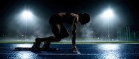 Athletic male runner in starting position on blue track under stadium lights preparing for sprint race in dramatic evening atmosphere.