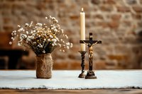 Burning candle in vintage brass candlestick with crucifix and rustic bouquet of white baby breath flowers in burlap vase on white tablecloth against stone wall.