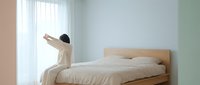 Woman in beige loungewear stretching arms above head while sitting on wooden bed in minimalist bedroom with natural light from window.