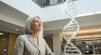 Senior Caucasian female scientist in beige suit examining large DNA double helix model in modern research laboratory or medical facility interior.