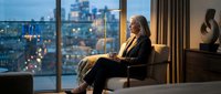 Woman in business attire sitting in modern office chair by floor-to-ceiling windows overlooking illuminated city skyline at dusk, suitable for corporate, professional, and lifestyle design projects.