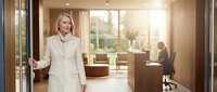 Professional blonde businesswoman in elegant beige suit walking through modern corporate office with warm lighting and contemporary furniture.