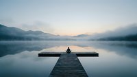 Person sitting alone on wooden dock at misty mountain lake during peaceful sunrise, perfect for meditation and mindfulness content.