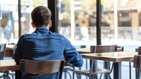 Young man in denim shirt sitting alone at empty cafe table looking through window, back view. Restaurant interior with wooden chairs and blurred city background.