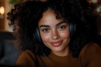 Young woman with dark curly hair and medium skin tone wearing black headphones and brown sweater, smiling at camera in warm indoor setting for music, audio, and lifestyle content.