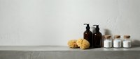 Minimalist bathroom shelf with natural sea sponge, dark amber glass bottles and white ceramic jars against textured wall for spa wellness routine.