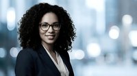 Young professional woman with dark curly hair and glasses wearing black blazer and white shirt, standing in modern office environment with blurred cityscape background.