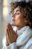Young African American woman with curly hair in white sweater and scarf praying peacefully with closed eyes and gentle smile in soft window light.