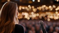 Young woman with long hair facing audience at event, microphone in foreground against blurred bokeh lights background.