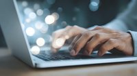 Close-up of male hands typing on laptop keyboard with soft bokeh lights in dark background, conveying late night work or creative inspiration.