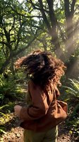 Young mixed-race woman with curly hair dancing in sunlit forest wearing earth-tone clothing surrounded by lush green trees and ferns.