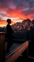 Woman in black clothing standing on wooden deck overlooking dramatic mountain landscape during vibrant sunset with fiery orange and red sky, suitable for travel and lifestyle content.
