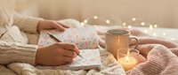 Woman writing in journal with warm candlelight and cozy knit blankets creating peaceful morning routine atmosphere for mindfulness and self-reflection.