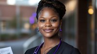 Young African American businesswoman with braided updo hairstyle wearing black blazer and purple accessories smiling confidently at camera in office setting.